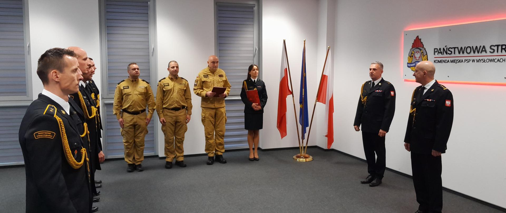 Strażacy stojący w pomieszczeniu sali sztabowej a naprzeciwko nich komendant miejski PSP wraz z zastępcą. W tle logo PSP i flagi Polski i UE