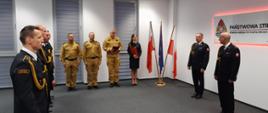 Strażacy stojący w pomieszczeniu sali sztabowej a naprzeciwko nich komendant miejski PSP wraz z zastępcą. W tle logo PSP i flagi Polski i UE