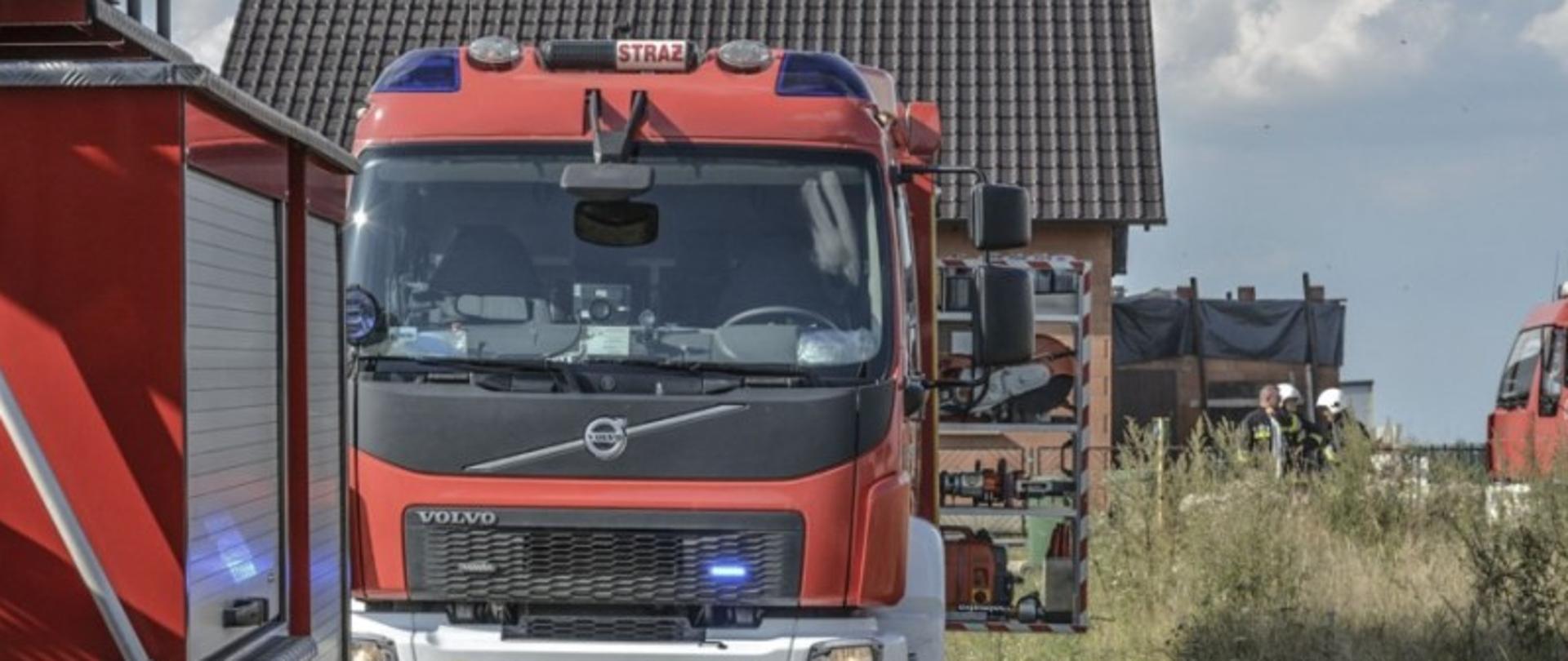 Na pierwszym planie fragment podnośnika hydraulicznego oraz średni samochód gaśniczy z Jednostki Ratowniczo - Gaśniczej w Krotoszynie. Pojazd ma włączone sygnały świetlne oraz otwartą skrytkę , gdzie znajdują się narzędzia ratownicze. W oddali zaparkowany samochód gaśniczy jednostki OSP oraz trzech druhów stojących przed nim. Za samochodami stoi budynek mieszkalny w trakcie budowy z brązowym spadzistym dachem. 