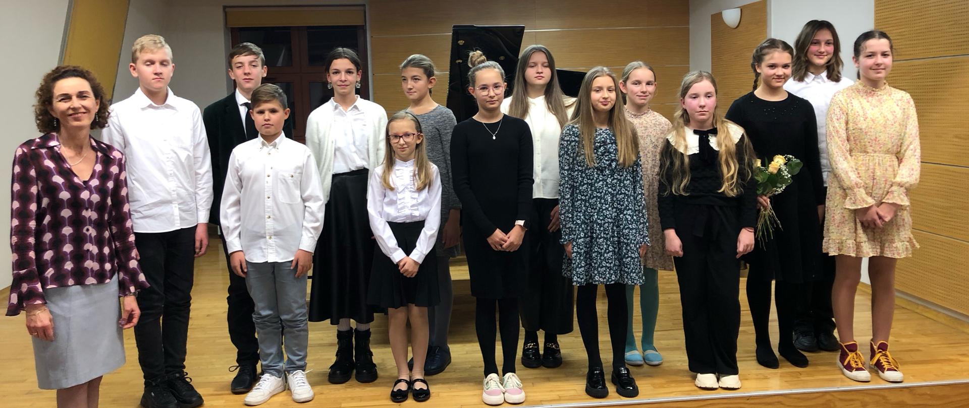 Młodzi pianiści stoją na scenie sali kameralnej w dwóch rzędach. z lewej strony stoi nauczycielka prowadząca audycje. Za nimi fortepiany, dwa okna przysłonięte roletą. nad nimi na suficie zapalone lampy sufitowe.