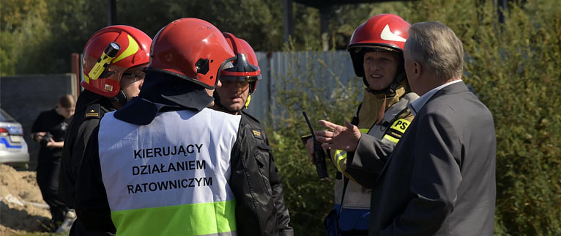 czterech strażaków rozmawia z osobą cywilna ubrana w garnitur, która ma uniesioną prawa dłoń lekko do góry, strażacy maja hełmy na głowie, strażak na pierwszy planie ma założoną kamizelkę z napisem „kierujący działaniem ratowniczym”, w tle widać zarośla, na dalszy planie metalowy płot z blachy przed którym stoi samochód osobowy i osoba z aparatem fotograficznym w dłoniach, obok tej osoby kupka piachu. Pogoda słoneczna