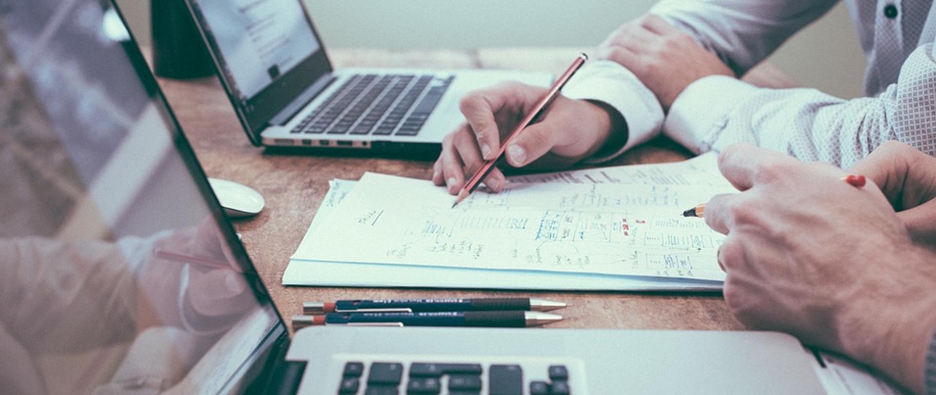 Dwa komputery, dokumenty, ręce trzymające ołówek i pisak