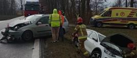 Na zdjęciu widać dwa rozbite samochody osobowe. Biały znajduje się w rowie. Ma otwartą maskę. Strażak w piaskowym stroju i czerwonym hełmie sprawdza coś w silniku. Srebrny pojazd stoi na drodze. Ma uszkodzony przód. Obok pojazdu stoi strażak w piaskowym stroju i czerwonym hełmie, a także dwie osoby. Za pojazdami widać żółtą karetkę pogotowia oraz czerwony samochód straży pożarnej. Obok pojazdów stoją policjanci i strażacy. W tle widać drzewa. Jest widno, ale pochmurnie.