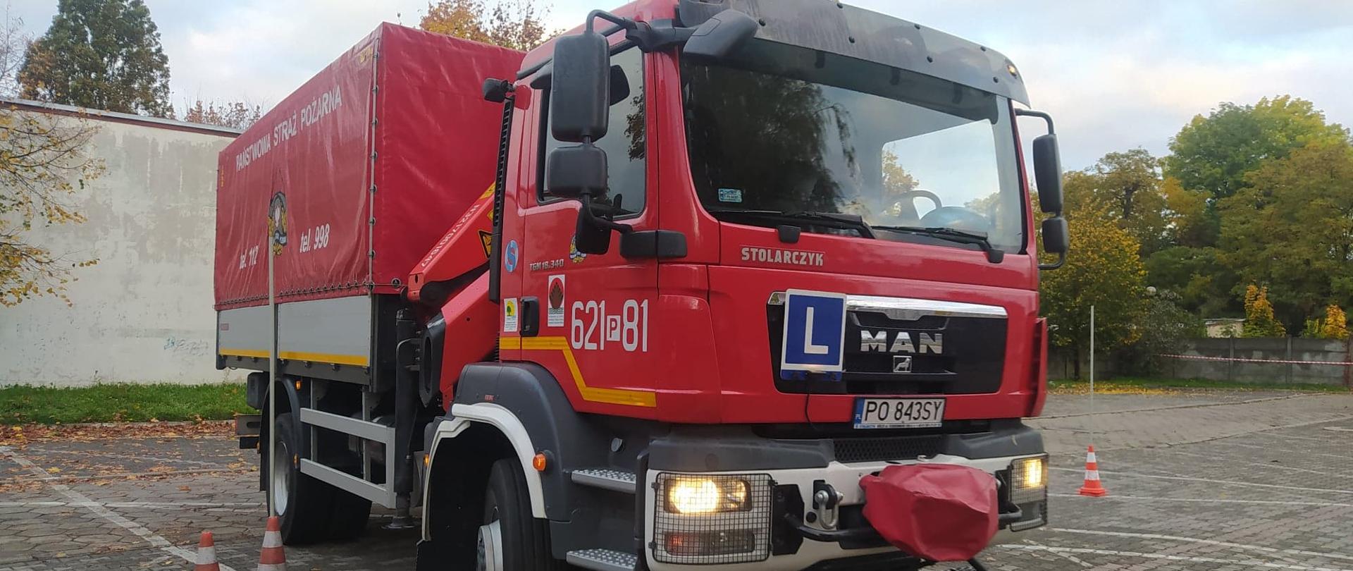 Zdjęcie przedstawia plac, na którym stoi wóz strażacki przeznaczony do nauki jazdy. Z przodu ma zawieszoną niebieską tabliczkę z białą literą "L" na środku. Samochód jest koloru czerwonego. . W tle budynek i drzewa.