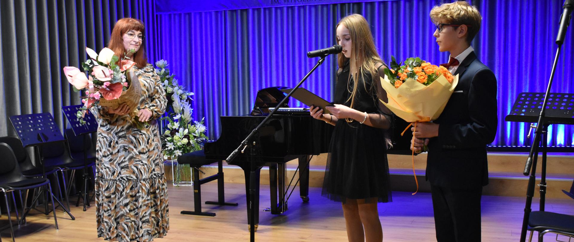 Na scenie auli PSM stoją przedstawiciele samorządu uczniowskiego, którzy kierują słowa podziękowania do stojącej obok Pani Dyrektor PSM. Pani Dyrektor trzyma w rękach duży bukiet kwiatów.