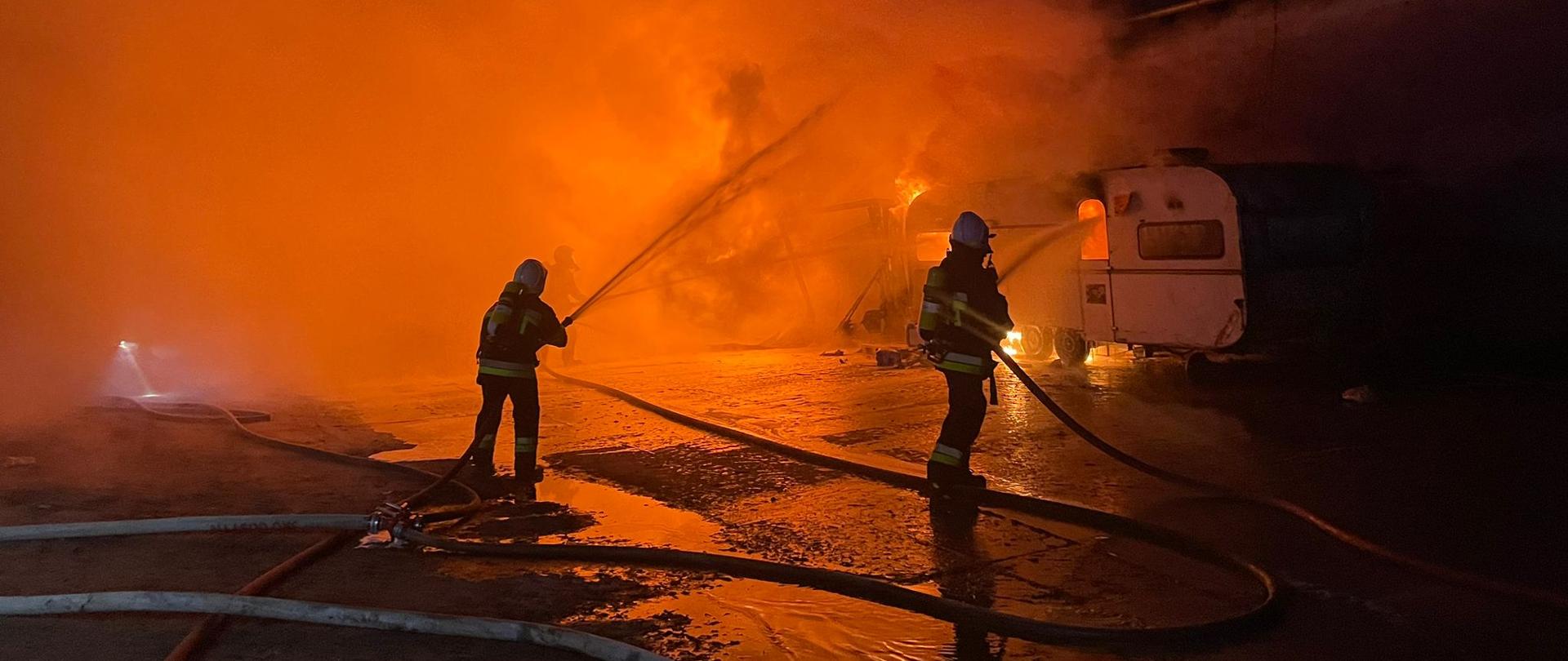 Dwóch strażaków gasi pożar zbiornika i przyczepy kempingowej.