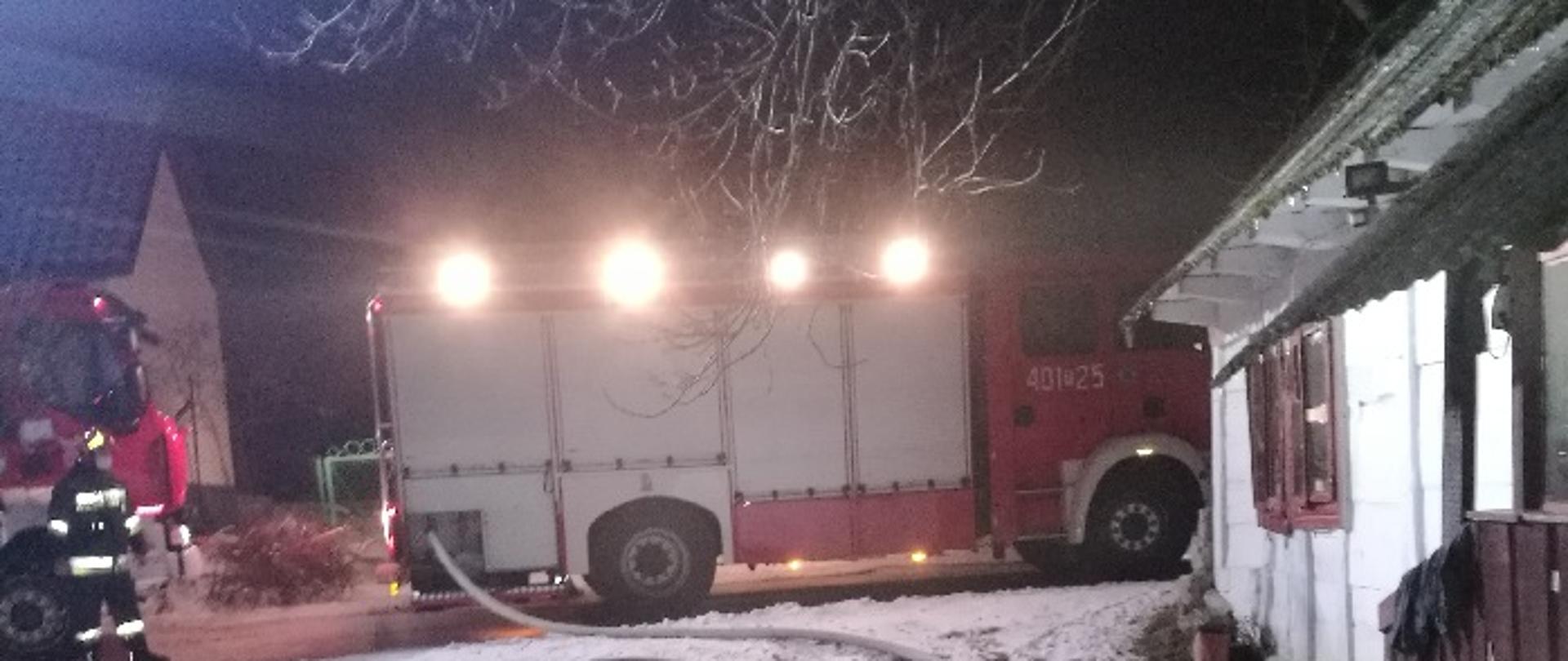 Zdjęcie przedstawia na wprost samochody pożarnicze, przy jednym stoi strażak w umundurowaniu specjalnym, do drugiego podpięte są węże gaśnicze, po prawej stronie dom parterowy drewniany pokryty eternitem ze ścianami w kolorze białym.