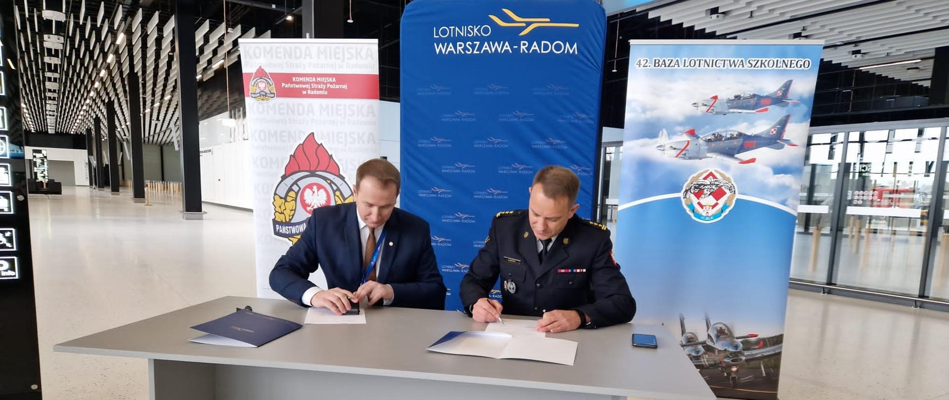 Zdjęcie wykonane na terenie portu lotniczego warszawa – radom podczas szkolenia kadry dowódczej z komendy miejskiej państwowej straży pożarnej w Radomiu. Na zdjęciu widoczny jest moment podpisania porozumienia pomiędzy dyrektorem lotniska warszawa – radom panem Grzegorzem Tuszyńskim, a komendantem miejskim państwowej straży pożarnej w Radomiu starszym brygadierem Bartoszem Musiałem, określające warunki współpracy państwowej straży pożarnej z lotniskową służbą ratowniczo – gaśniczą.