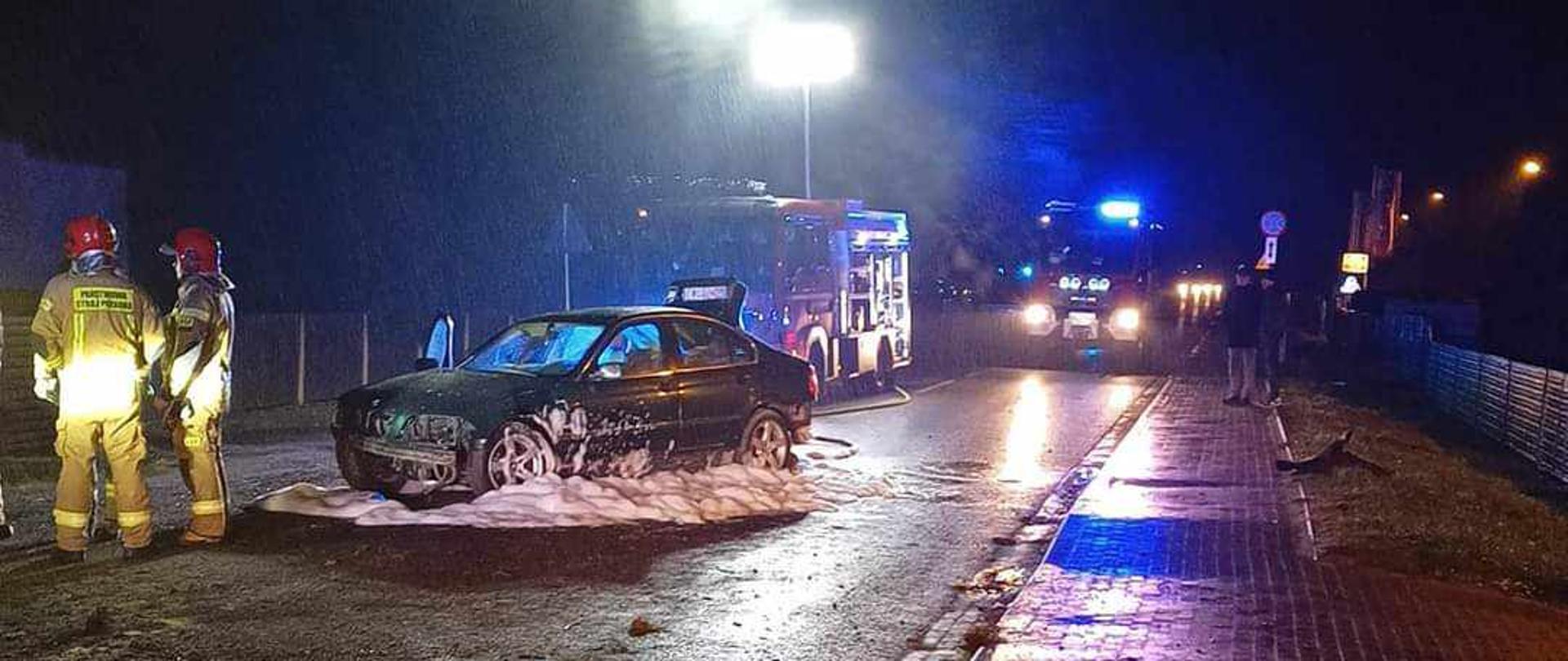 Zdjęcie przedstawia samochód osobowy stojący na jezdni. Po lewej stronie stoi dwóch strażaków. Za samochodem osobowym stoją dwa wozy straży pożarnej
