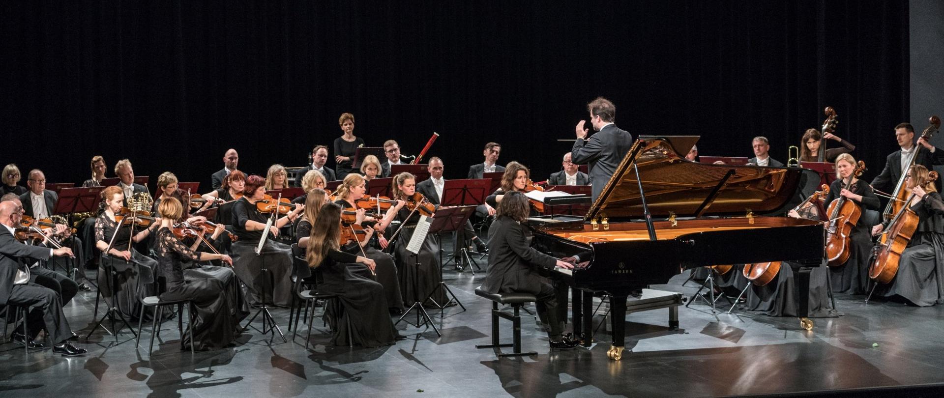 Orkiestra siedzi na scenie, pianista przy fortepianie, dyrygent stoi przodem do orkiestry