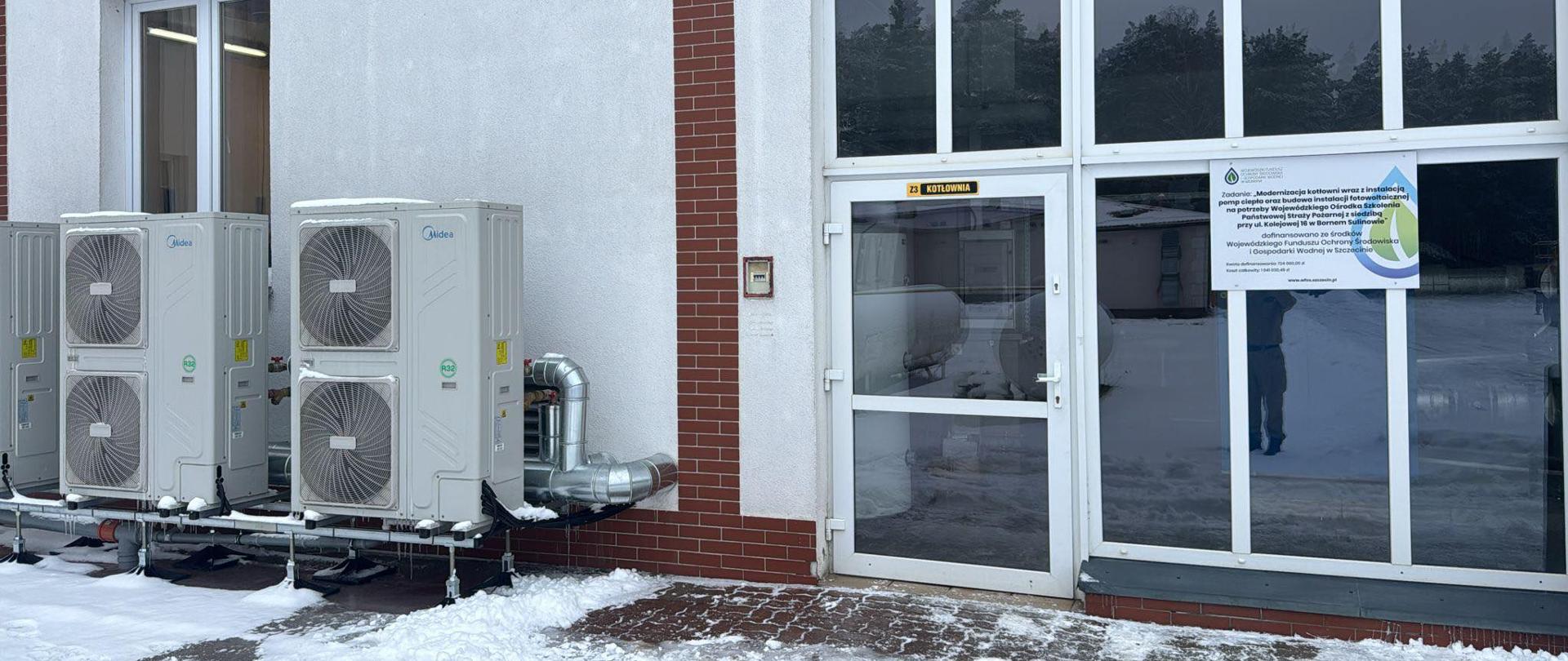 Zdjęcie przedstawia elewację budynku zimą. Po lewej stronie znajdują się cztery zewnętrzne jednostki pomp ciepła, a po prawej przeszklone wejście z napisem „Kotłownia” i plakatem informacyjnym. Przed budynkiem widać śnieg i oblodzony chodnik.