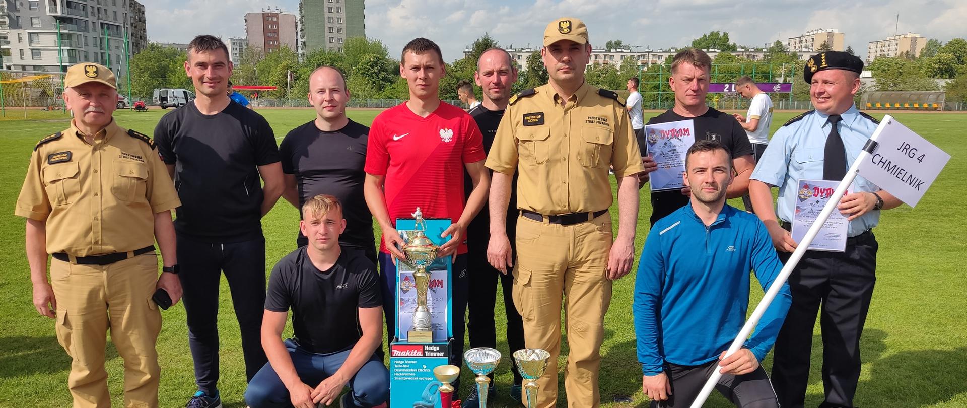 Zdjęcie prezentuje drużynę sportowców z Jednostki Ratowniczo-Gaśniczej nr 4 w Chmielniku w towarzystwie Świętokrzyskiego Komendanta Wojewódzkiego PSP nadbryg. Krzysztofa Cioska oraz Zastępcy Świętokrzyskiego Komendanta Wojewódzkiego PSP st. bryg. Jarosława Piotrowskiego. 