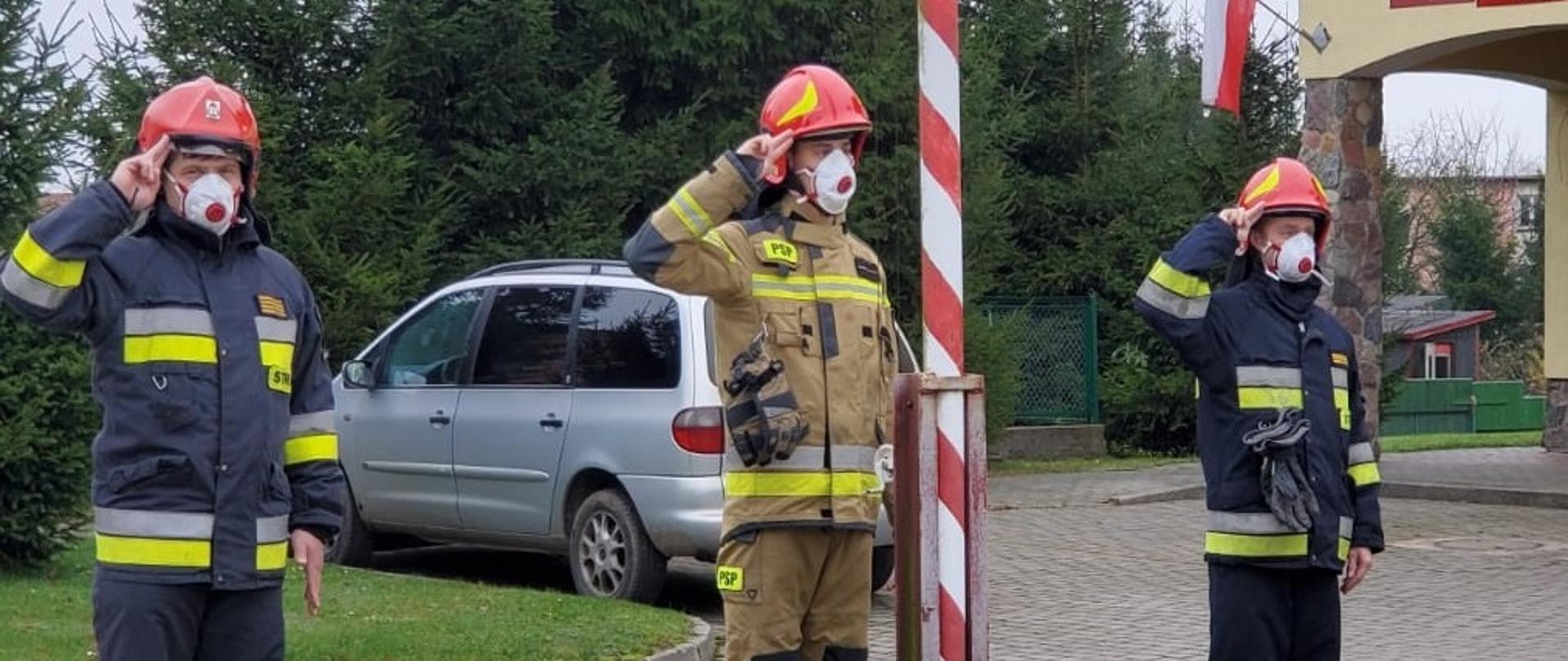 Trzech strażaków ubranych w ubranie specjalne typu NOMEX stojących na baczność przy maszcie, oddających honor do flagi państwowej. W oddali widać smaochód osobowy vw sharan oraz żółty budynek komendy. 