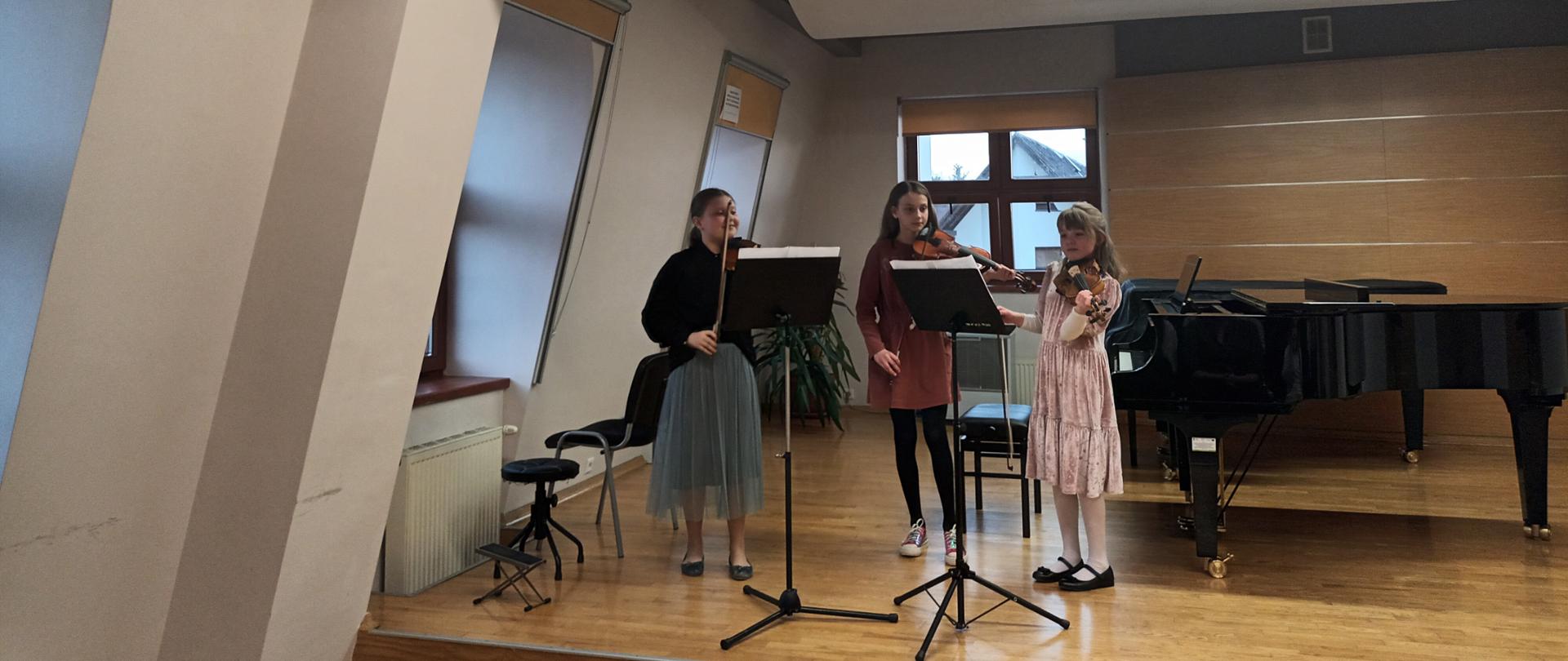 Na sali kameralnej na scenie stoją trzy skrzypaczki, trzymają skrzypce gotowe do grania, przed nimi dwa pulpity, a na nich nuty. Z lewej strony podnóżek do gitary, taboret i krzesło, okno oraz kaloryfer. Za nimi dwa fortepiany, ława fortepianowa, nawilżacz oraz okno.