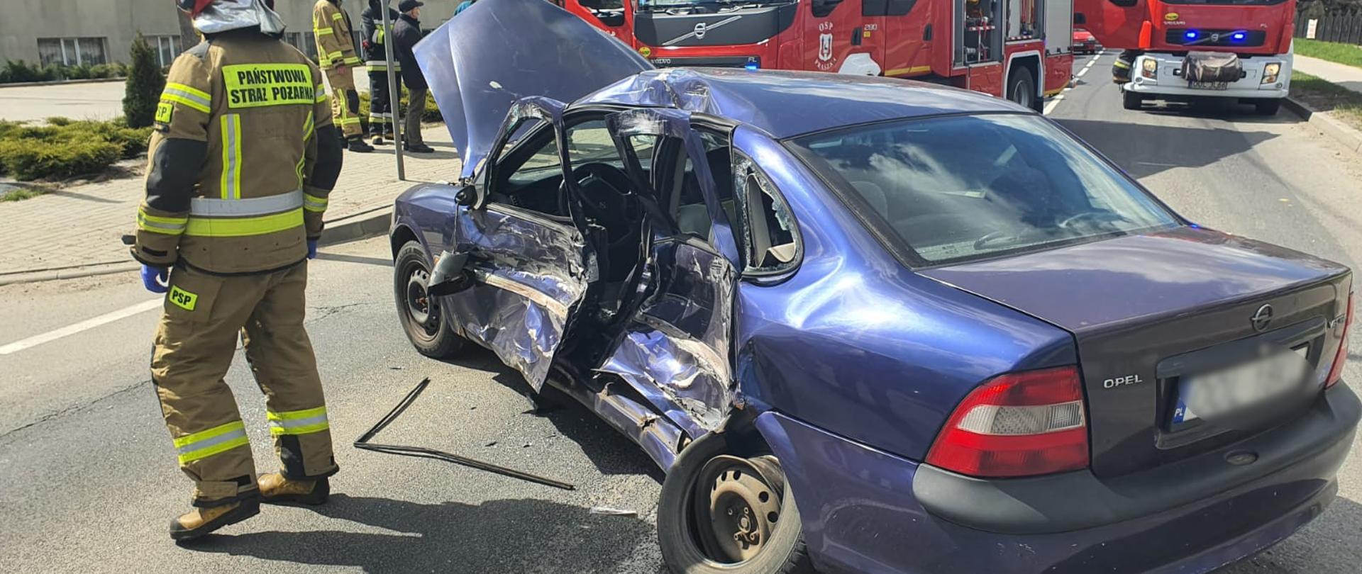 Na zdjęciu samochód osobowy koloru fioletowego stoi w poprzek drogi z uszkodzonym lewym bokiem. Obok samochodu strażak w ubraniu specjalnym koloru piaskowego oraz czerwonym hełmem. Z tył samochodu stoją strażacy oraz dwa samochody pożarnicze.