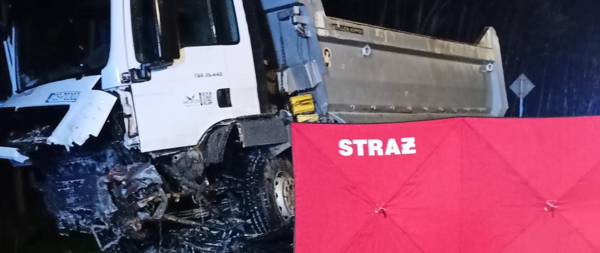 Zdjęcie przedstawia stojący na poboczu samochód ciężarowy ze zniszczonym przodem, pod którym znajduje się fragment samochodu osobowego. Przestrzeń przy ciężarówce osłonięta jest parawanem straży pożarnej.