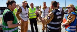 Na zdjęciu widoczna jest grupa osób uczestniczących w organizacji oraz nadzorowaniu przebiegu ćwiczeń. Uczestnicy wyposażeni są w kamizelki funkcyjne umożliwiające identyfikację pełnionych przez nich ról. W centrum kadru prowadzona jest rozmowa dotycząca realizowanych działań, a uczestnicy wymieniają informacje związane z przebiegiem ćwiczeń.
Fotografia obrazuje istotny element przedsięwzięcia, jakim jest bieżąca obserwacja działań poszczególnych zespołów, wymiana informacji oraz ocena sposobu realizacji założonych zadań. Obecność osób pełniących funkcje obserwacyjne pozwala na późniejszą analizę przebiegu ćwiczeń i ocenę współdziałania zaangażowanych podmiotów.