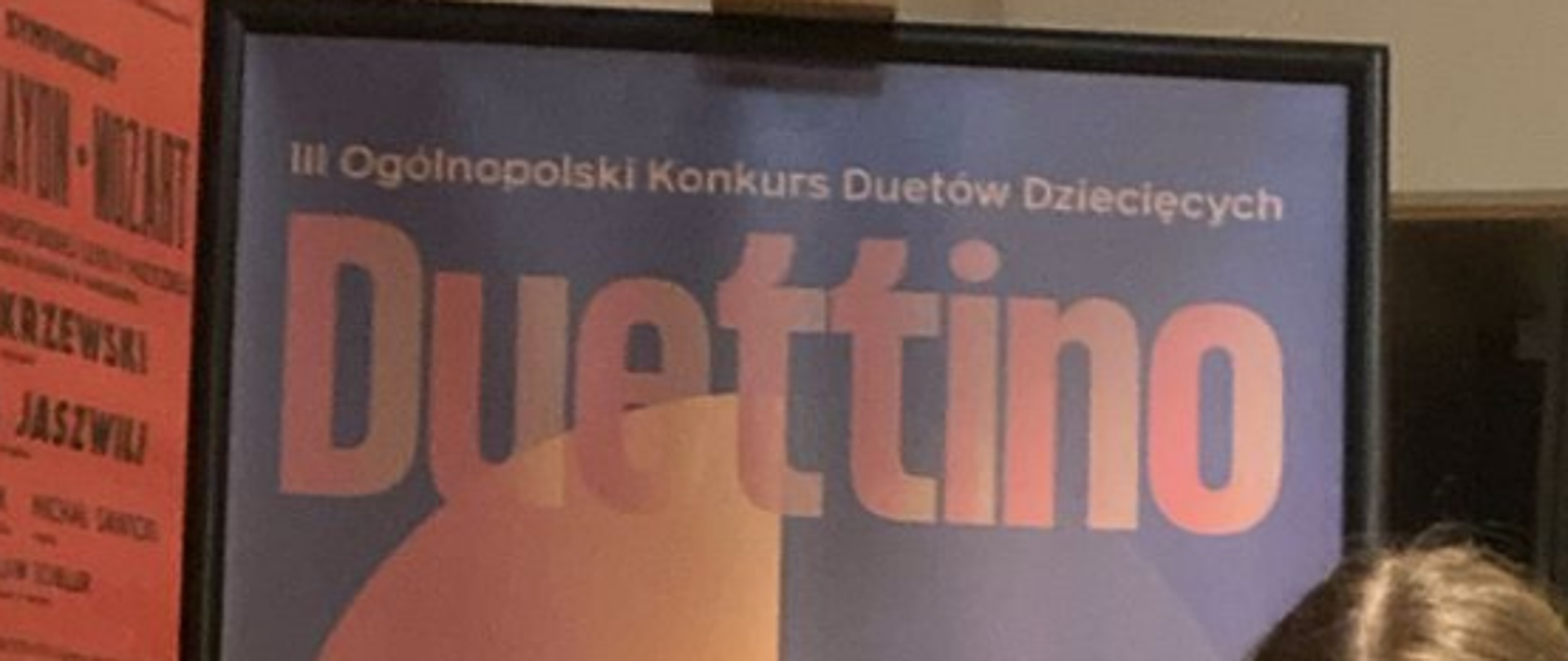 Zdjęcie przedstawia 4 uczniów (1 chłopiec i 3 dziewczynki) szkoły trzymających dyplomy wyróżnienia na Konkurskie Duettino. Ubrani odświętnie, 2 dziewczynki mają spódnice z materiału we wzór zapisów nutowych. W tle widać afisz konkursowy, Na dole zdjęcia zawarta jest informacja o zdobytych wyróżnieniach duetów: Józefina Jurdzińska - flet i Maria Mila - fortepian oraz Estera Matlak - flet i Aleksander Matlak - trąbka.
