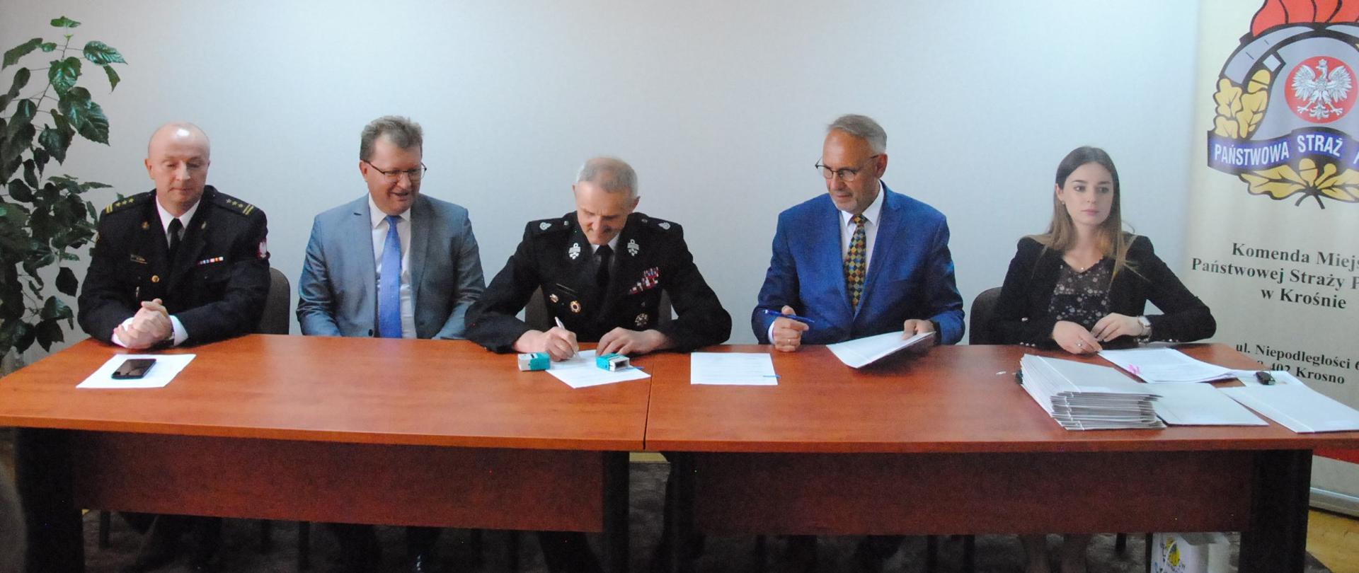 Zdjęcie przedstawia 5 osób siedzących za stołem prezydialnym w Sali konferencyjnej KM PSP Krosno składających podpisy pod umowami na dofinansowanie zakupu sprzętu dla jednostek OSP. Za siedzącymi, na białej ścianie widoczne jest godło Polski a z prawe strony logo Państwowej Straży Pożarnej w Krośnie.