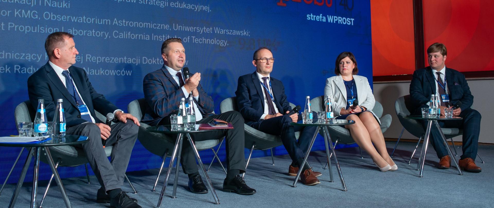 Czterech mężczyzn w garniturach i ubrana na szaro kobieta siedzą na ustawionych w rzędzie krzesłach, przed nimi małe stoliki, za nimi niebieska ściana.