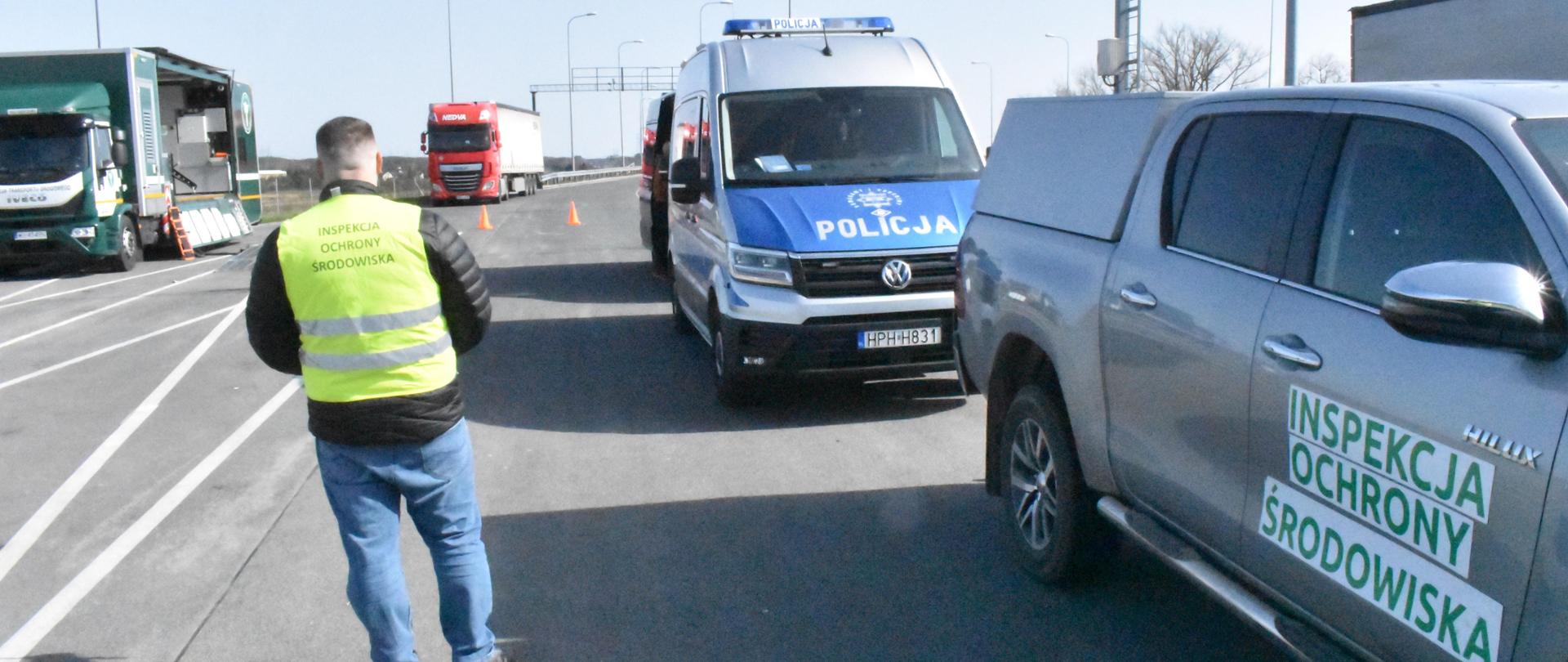 Inspektor Wojewódzkiego Inspektoratu Ochrony Środowiska w Warszawie prowadzi działania w punkcie kontrolnym na drodze. Po jego prawej stronie stoi samochód osobowy należący do Inspekcji Ochrony środowiska oraz furgonetka należąca do Policji. W tle widać dwa samochody ciężarowe.
