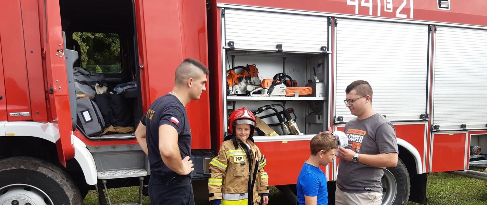 Na zdjęciu widać mężczyznę i dwójkę dzieci. Jedno z dzieci ma założoną żółtą kurtkę strażacką i czerwony hełm. Obok nich stoi strażak w czarnym stroju. W tle widać czerwony samochód strażacki, który ma otwarte drzwi i skrytki ze sprzętem. Na boku ma biały napis 441E21. Jest widno