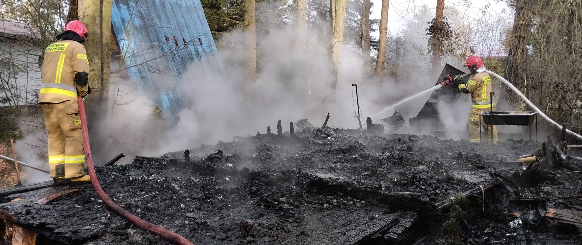 Na zdjęciu widać jak dwóch strażaków dogasza zarzewia ognia po pożarze budynku letniskowego typu tramp. W tle widać drzewa oraz inny budynek letniskowy