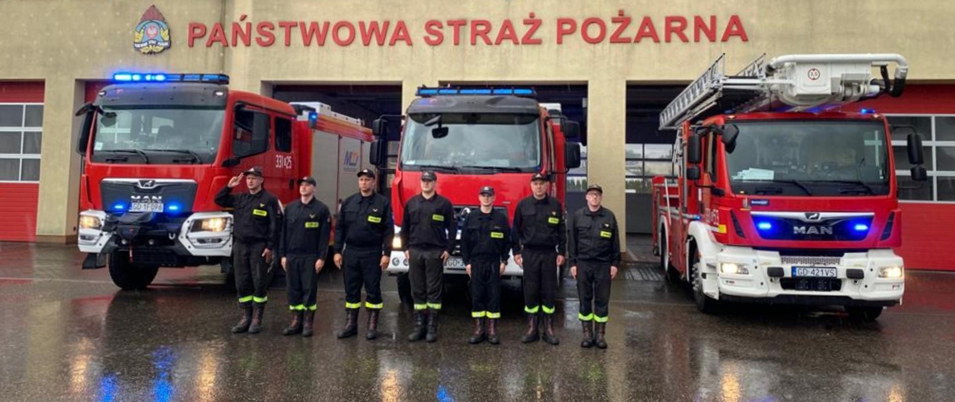 Zdjęcie przedstawia funkcjonariuszy JRG w Pruszczu Gdańskim oddających hołd Powstańcom Warszawskim na tle samochodów pożarniczych z włączoną sygnalizacją świetlno-dźwiękową.