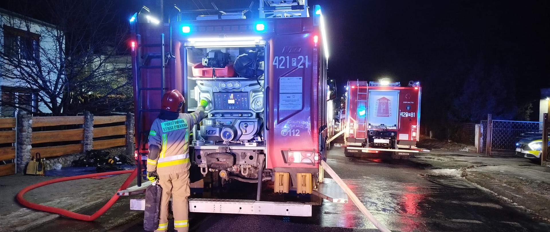 Jest pora nocna, na zdjęciu widać samochody pożarnicze. Kierowca, który stoi za samochodem pożarniczym obsługuje autopompę podając w ten sposób wodę poprzez węże pożarnicze, która jest wykorzystywana podczas gaszenia pożaru.