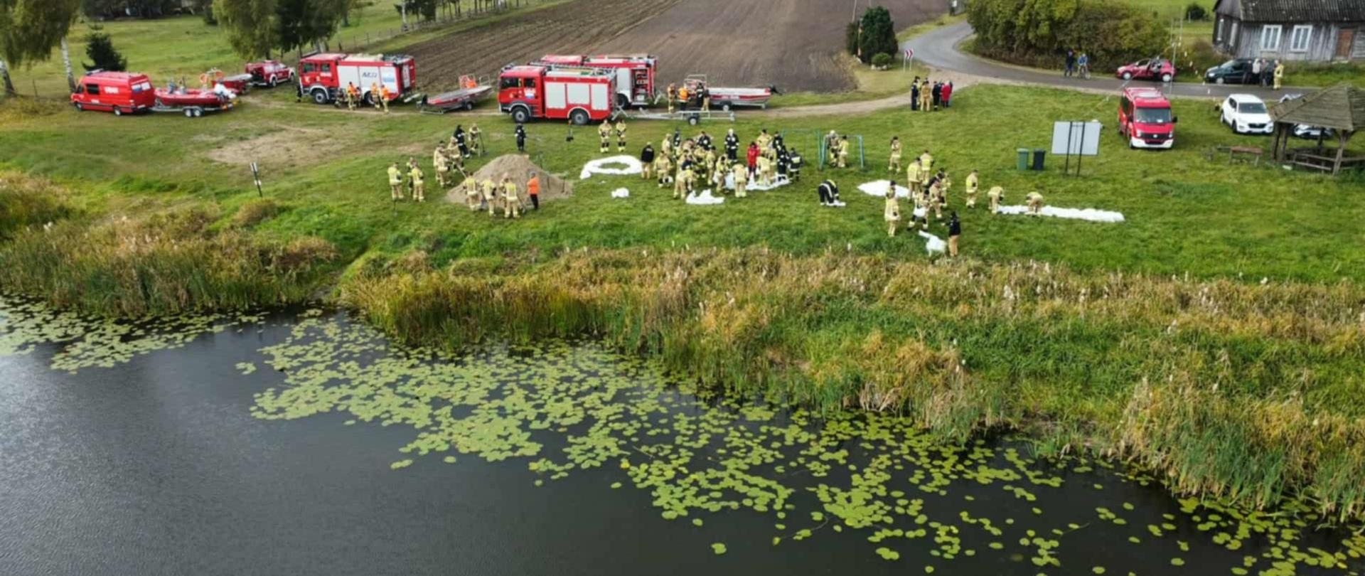 Na zdjęciu widzimy dużą grupę strażaków na trawiastym terenie przy zbiorniku wodnym. Większość osób ubrana jest w mundury w kolorze beżowym lub żółto-beżowym, które wydają się być odzieżą roboczą lub ochronną, charakterystyczną dla służb ratunkowych lub straży pożarnej. Kilka osób w środku grupy ma ciemniejsze, granatowe lub czarne ubrania. Po lewej stronie widać też jedną osobę w czerwonej kurtce z odblaskowymi elementami. Wszyscy są zwróceni w kierunku obiektywu. W tle, za grupą ludzi, widać linię drzew i krzewów, a niebo jest szare i zachmurzone.