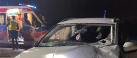 Na zdjęciu znajduje się wrak samochodu osobowego, w tle widać karetkę pogotowia oraz stojących przy niej ludzi