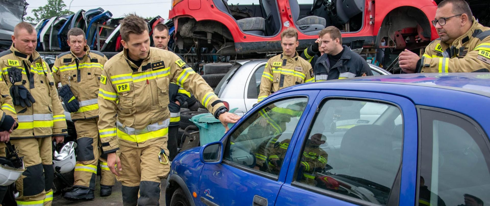 Zdjęcie przedstawia strażaka stojącego przy samochodzie, który jest przygotowany do ćwiczeń. na drugim planie widać 7 strażaków przysłuchujących się instruktorowi. W tle rozebrane auta i ich części.