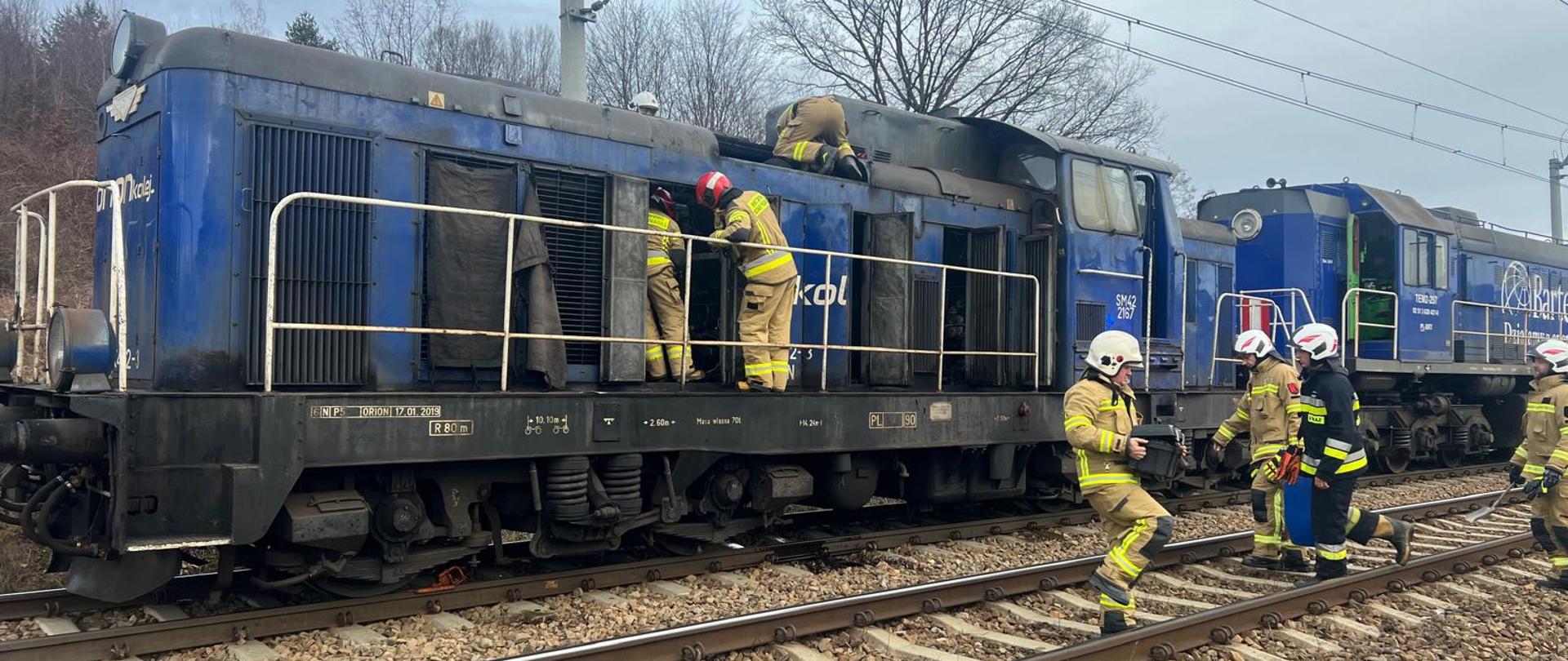 Dwie lokomotywy na torach i strażacy prowadzący działania