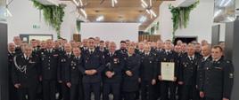 Na zdjęciu widać dużą grupę osób ubranych w uroczyste mundury strażackie, stojących razem w eleganckim, nowoczesnym wnętrzu. W centralnej części znajdują się osoby wyróżnione, a jedna z nich trzyma w rękach duży, oprawiony dyplom lub akt nadania.