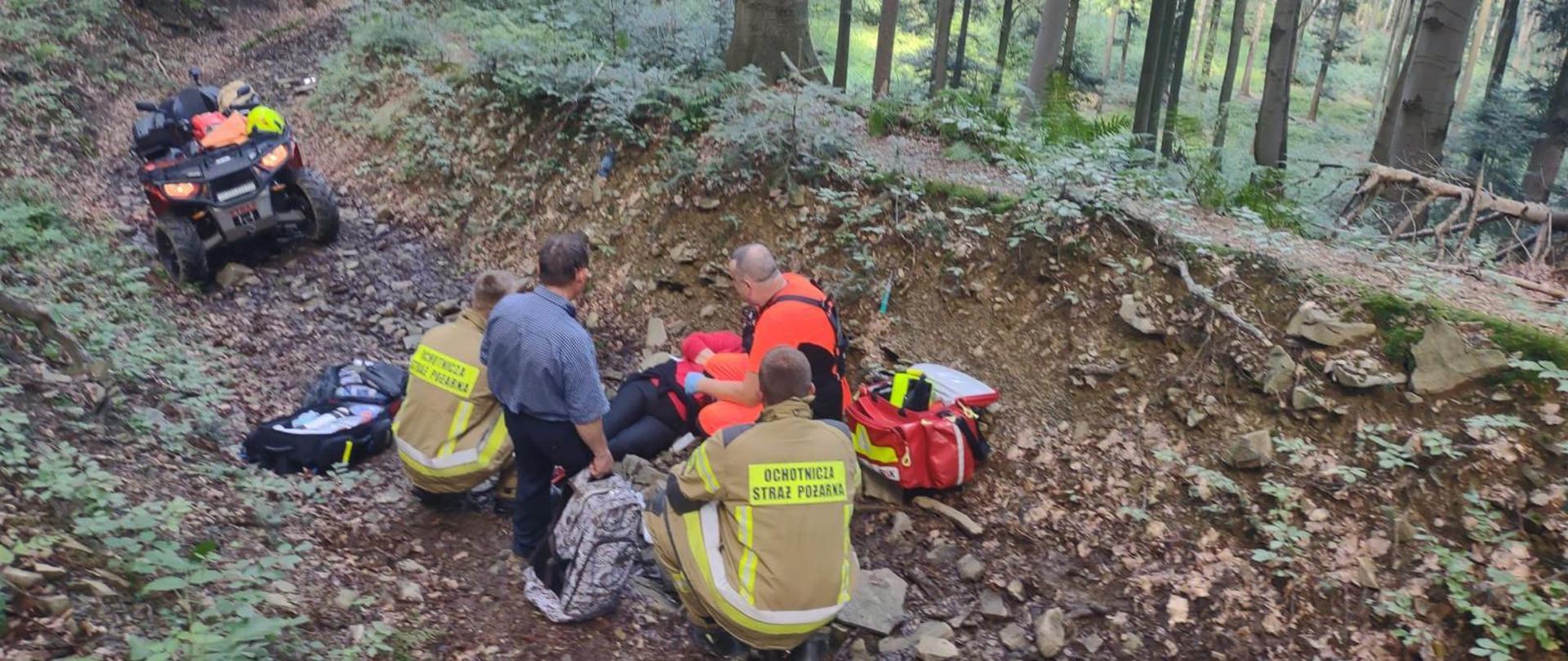 Zdjęcie przedstawia strażaków wraz z ratownikiem medycznym, którzy udzielają pomocy poszkodowanej turystce. Turystka doznała urazu na szlaku turystycznym na Gorę Kamionna. W tle stoi Quad którym turystka będzie ewakuowana po udzieleniu pierwszej pomocy.