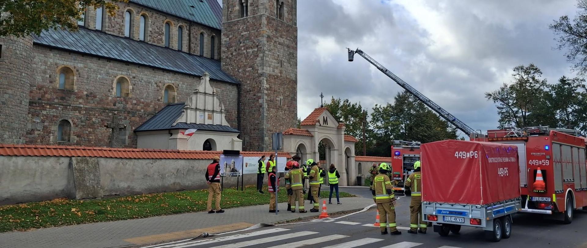 Na zdjęciu widać strażaków zgromadzonych przed zabytkowym kościołem oraz samochody pożarnicze zaparkowane wzdłuż drogi. Jeden z pojazdów ma rozłożony podnośnik hydrauliczny, a teren działań jest zabezpieczony pachołkami.