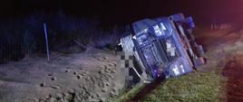 Kolizja na drodze krajowej. Auto ciężarowe leży w rowie na swoim prawym boku. Wokół rozsypany jest materiał przewożony pojazdem (kruszywo).