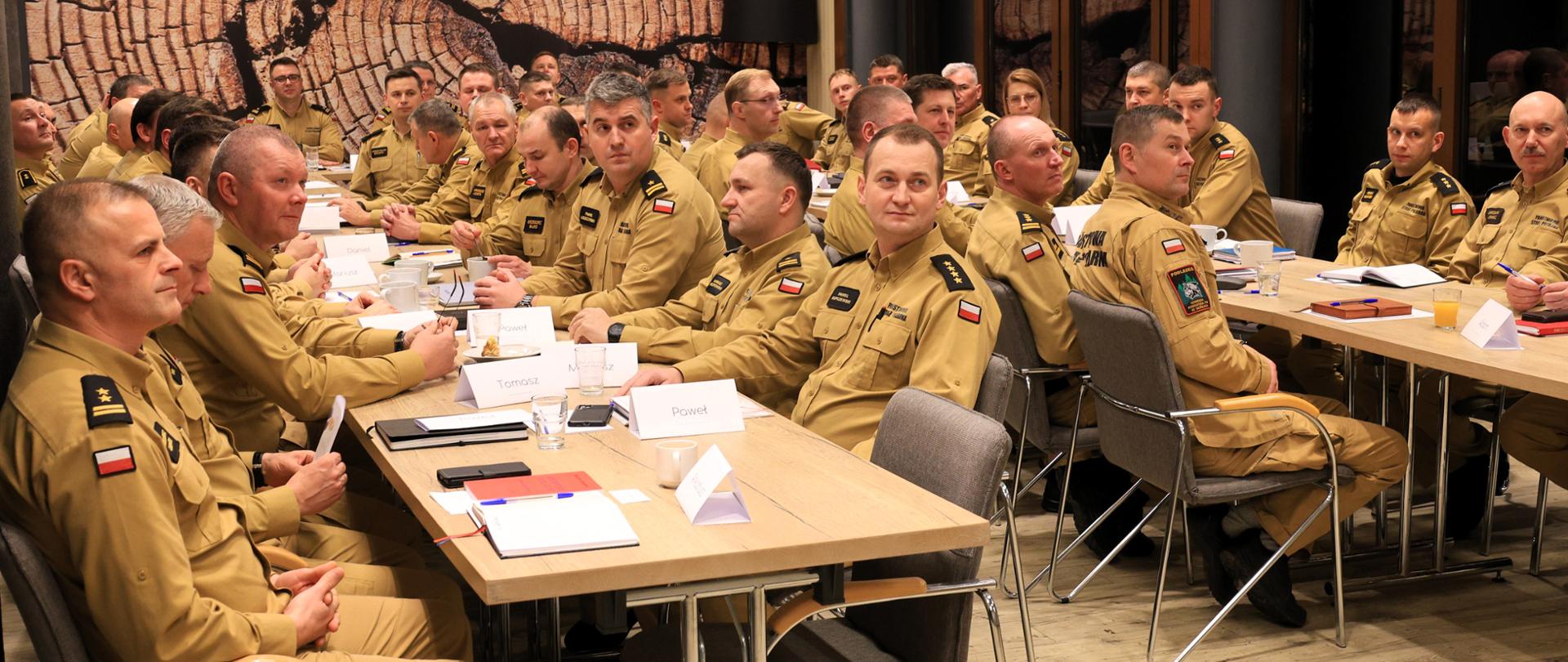 Odprawa szkoleniowa skierowana do kadry kierowniczej, a także oficerów prasowych z terenu województwa podlaskiego - strażacy siedzą przy stołach ustawionych w kształt litery "U", podczas zajęć wykładowych, na sali szkoleniowej.