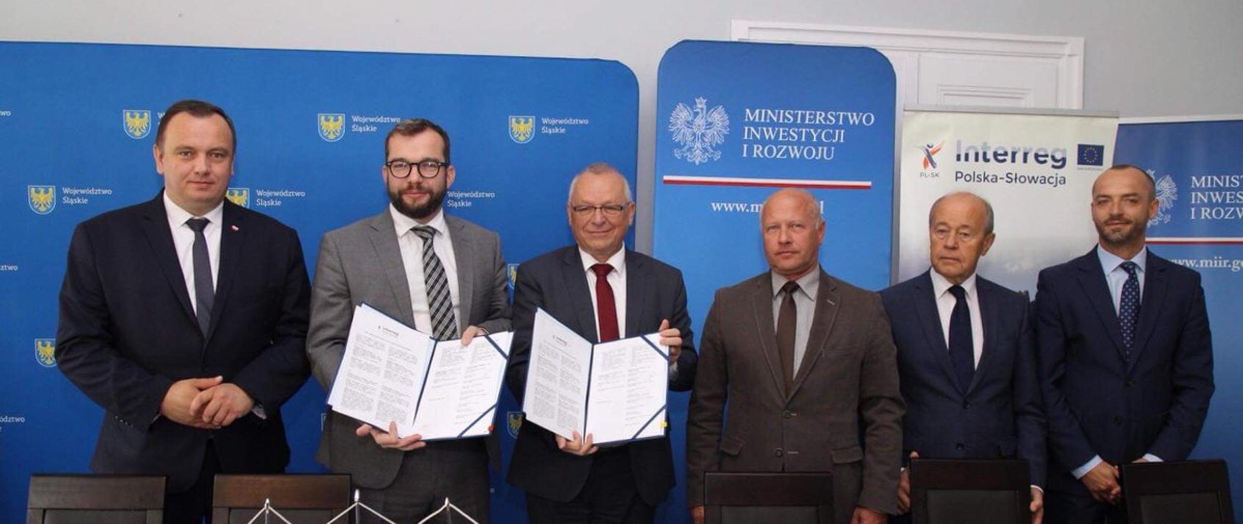 Minister Grzegorz Puda stoi i trzyma w ręku podpisaną umowę. Obok niego stoi pięciu pozostałych uczestników spotkania. 