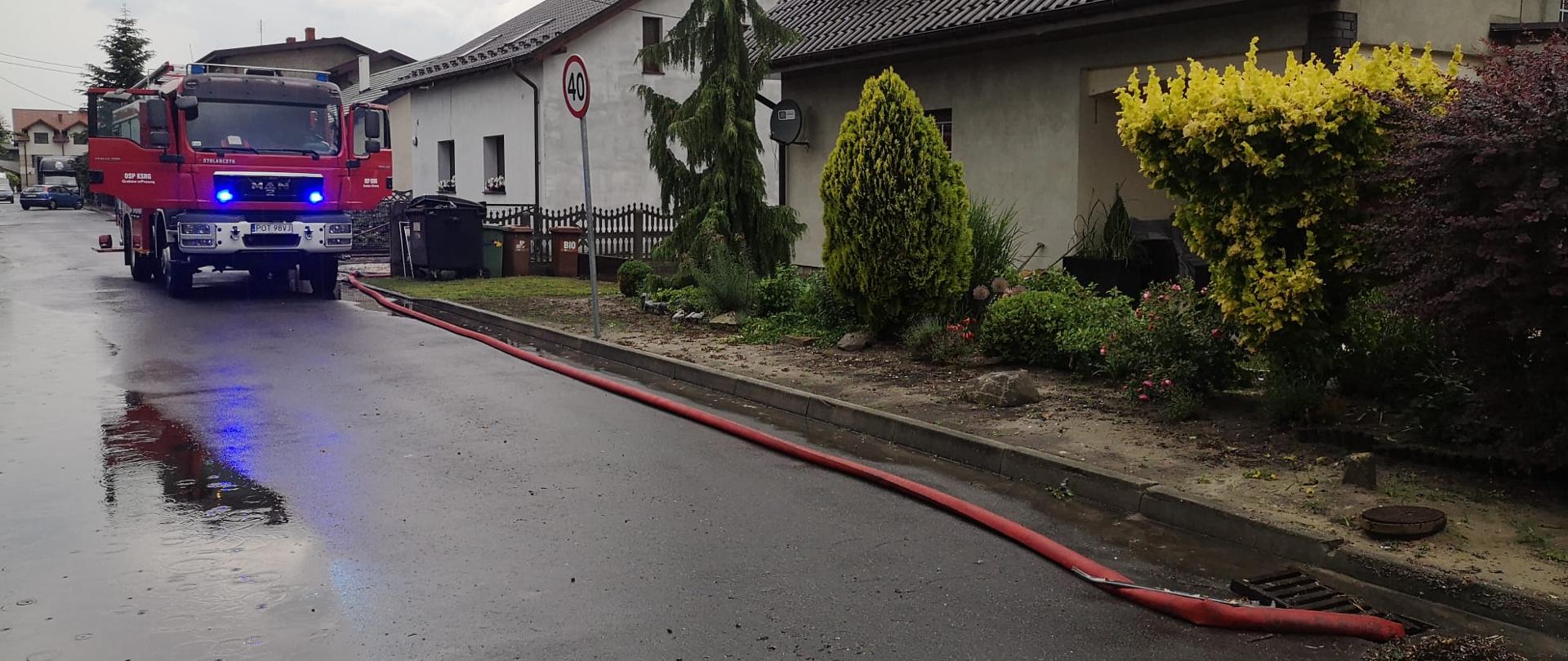 Samochód strażacki stoi na drodze , wzdłuż której rozłożony jest wąż odprowadzający wodę z zalanej posesji. Po prawej stronie budynek mieszkalny, a przed nim krzewy ozdobne.