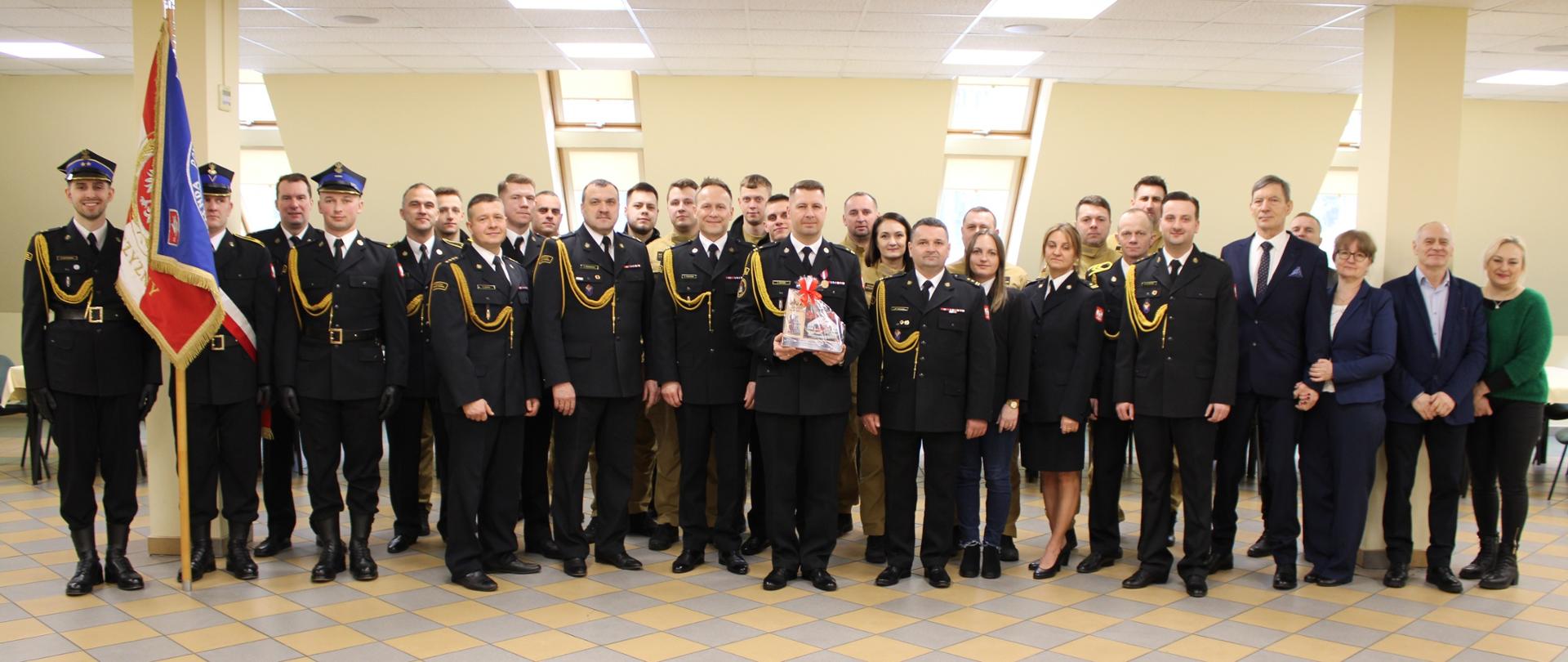 Uczestnicy uroczystej zbiórki stoją w mundurach i pozują do zdjęcia z okazji zakończenia służby w Komendzie Powiatowej Państwowej Straży Pożarnej w Golubiu-Dobrzyniu przez mł. bryg. Łukasza Buczka.