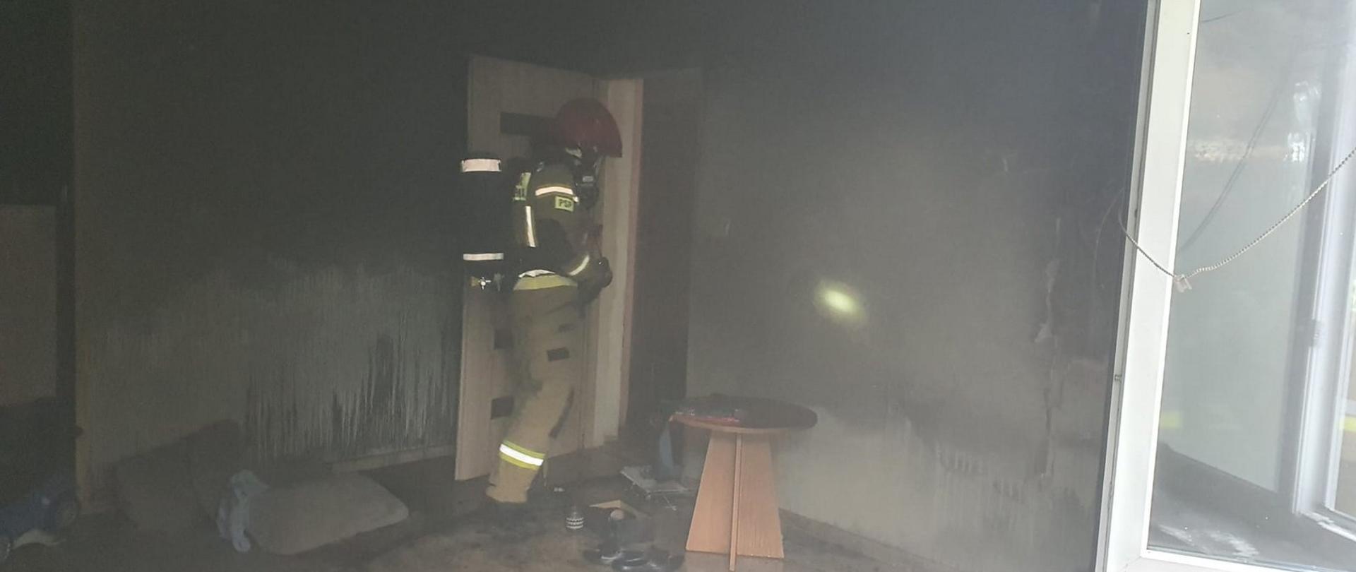 Zdjęcie zrobione w pokoju objętym pożarem. Pomieszczenie z obkopconymi ścianami. Wewnątrz strażak ubrany w piaskowe ubranie specjalne, czerwony hełm oraz aparatem ochrony układu oddechowego.