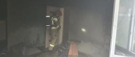 Zdjęcie zrobione w pokoju objętym pożarem. Pomieszczenie z obkopconymi ścianami. Wewnątrz strażak ubrany w piaskowe ubranie specjalne, czerwony hełm oraz aparatem ochrony układu oddechowego.