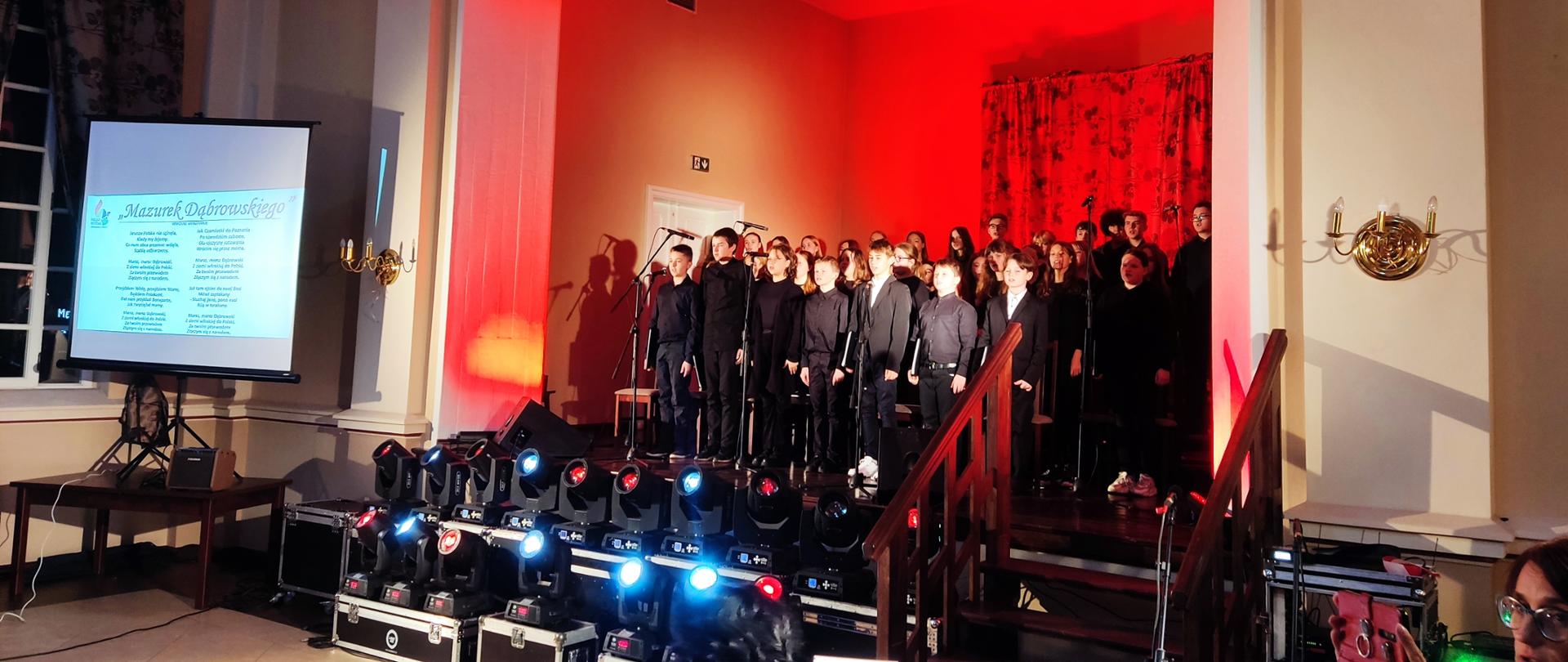 Zdjęcie przedstawia stojących na scenie w kilku rzędach i śpiewających chórzystów. Chórzyści ubrani są na czarno. Na scenę rzucone jest czerwone światło, stanowiące tło. Za chórem widoczna jest zasunięta kotara w kwiatowy wzór. Przed chórem ustawione są mikrofony na statywach. Przed sceną w dwurzędzie (niżej i wyżej) ustawione są światła, skierowane w stronę publiczności, świecące naprzemiennie na niebiesko i czerwono. Z prawej strony widoczne są drewniane schody z poręczami prowadzące na scenę. Z lewej strony na brązowym stole ustawiony jest na statywie i rozwinięty ekran, na którym wyświetlony jest tekst Mazurka Dąbrowskiego. 