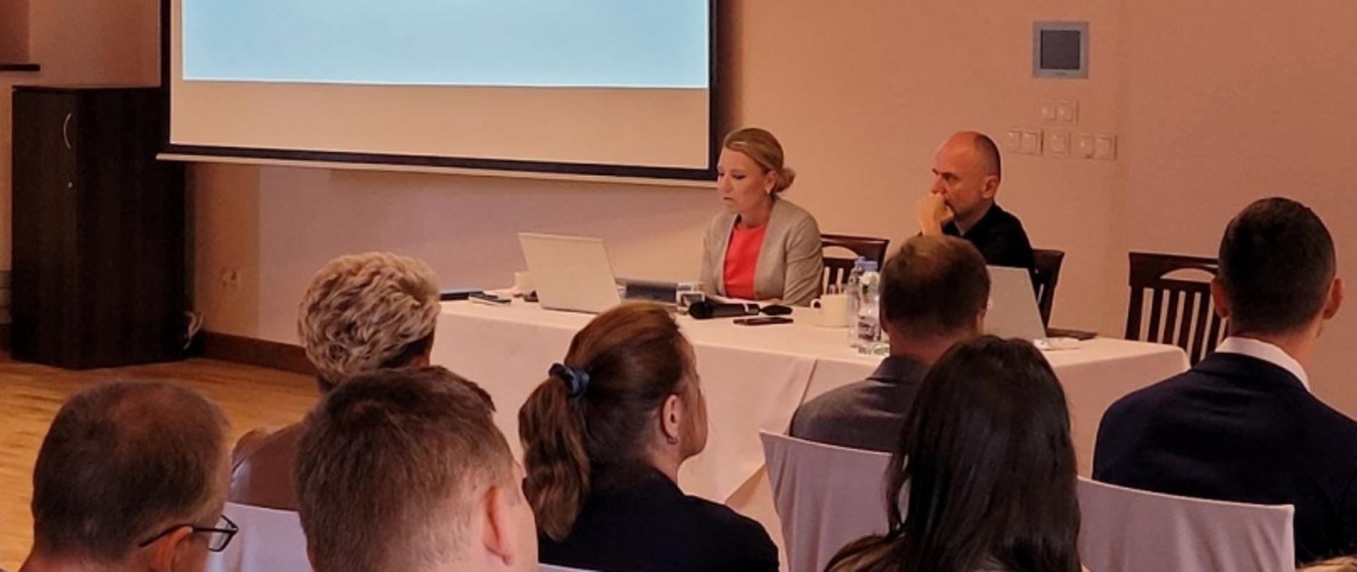 Spotkanie lub prezentacja z udziałem publiczności; dwóch prelegentów siedzi przy stole z laptopami i mikrofonami.