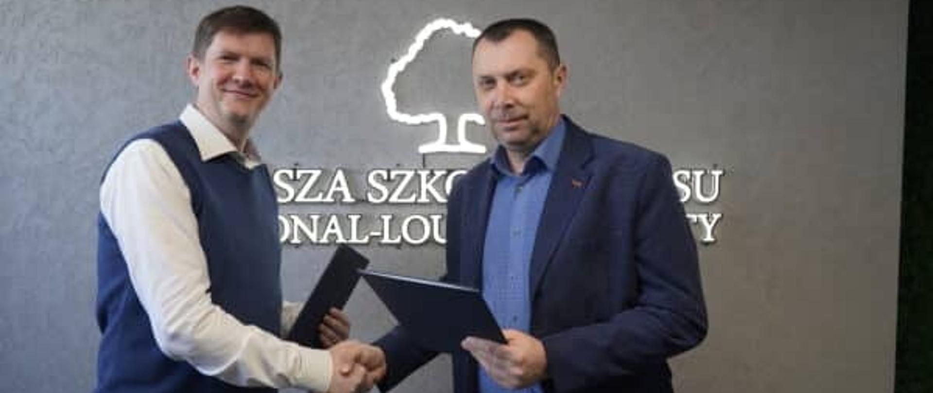 Dwaj mężczyźni uśmiechają się i podają sobie ręce, trzymając podpisane teczki z dokumentami na tle logo szkoły.