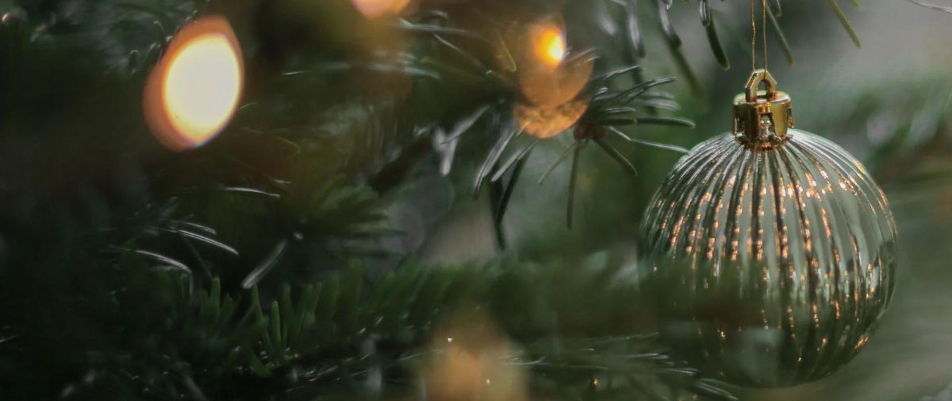Niewyraźne zdjęcie gałązki choinkowej. Na niej wyostrzony obraz zawieszonej okrągłej bombki koloru złotosrebrnego. Widoczne są również rozmyte światełka.