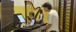 Zdjęcie przedstawia troje młodych muzyków, pianistę, perkusistę i harfistkę, podczas próby orkiestry, widać instrumenty perkusyjne, harfę, elementy wystroju szkolnego 