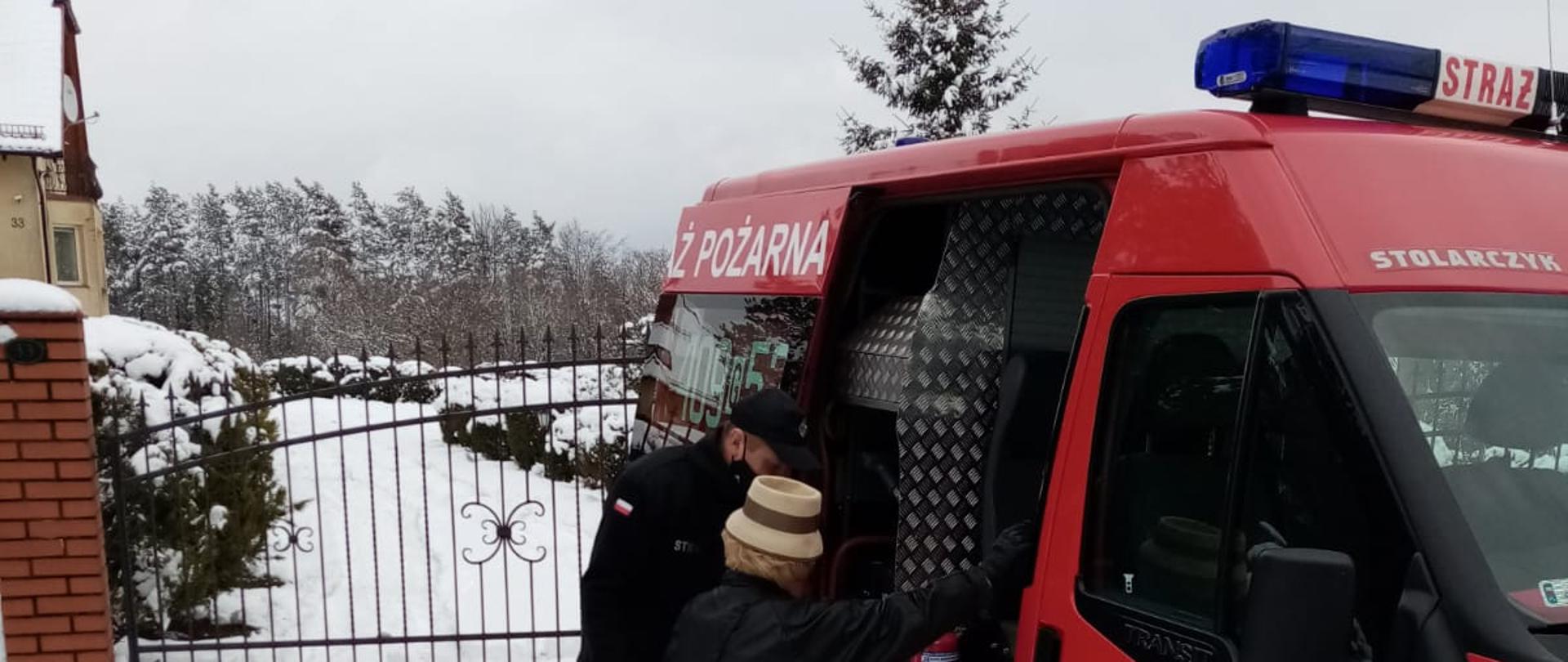 Zdjęcie transportu osób do punktu szczepień. Zdjęcie przedstawia strażaka w ubraniu koszarowym koloru czarnego pomagającego wysiąść osobie starczej z samochodu pożarniczego