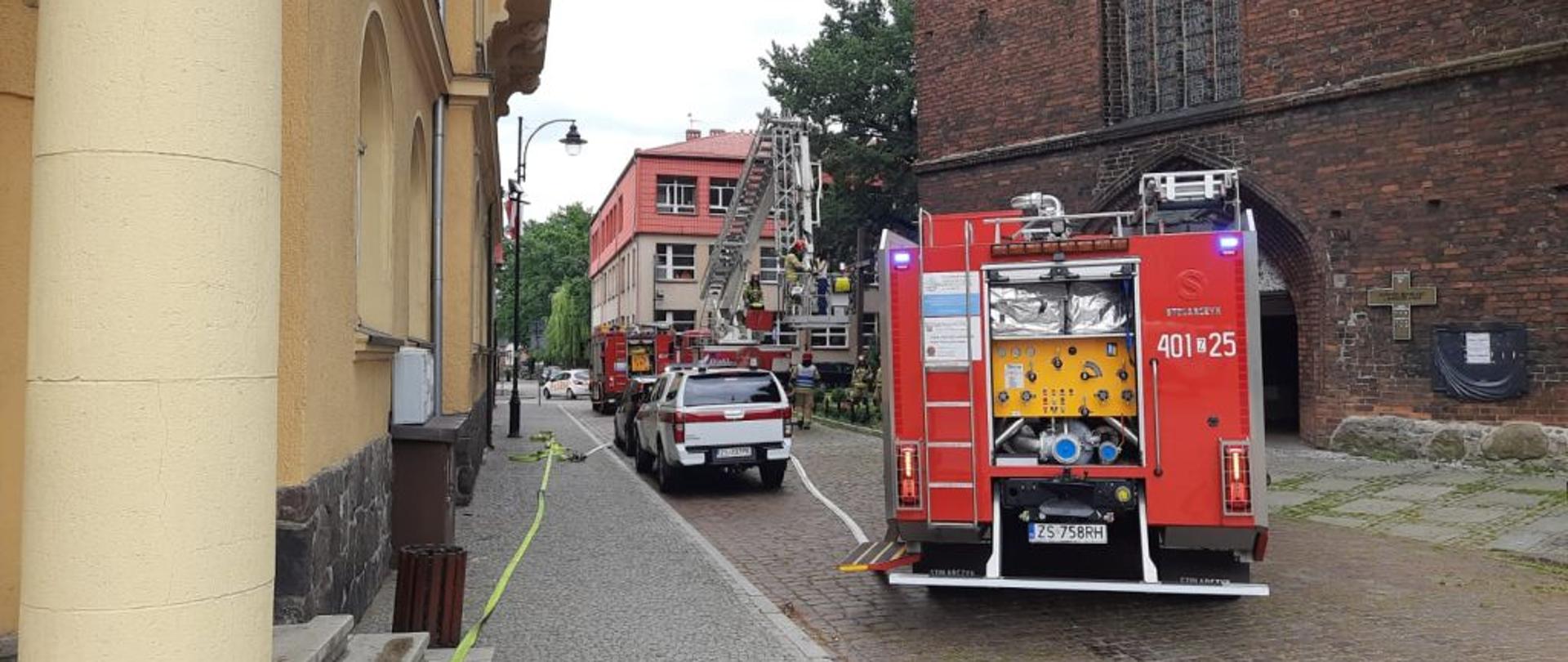 Widok na stojące na ulicy wozy strażackie. W tle budynek kościoła. Po lewej żółty budynek starostwa. Jest ładny pogodny dzień.
